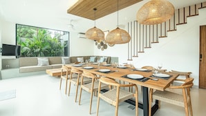 A modern dining area with a wooden table set for six, surrounded by wicker chairs. A staircase with wooden steps and a white wall is visible in the background. - Chic 5BR Canggu Villa 400m to Café Del Mar (Seminyak)