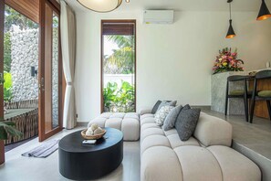 A modern living room with a beige sofa, a black round coffee table, and a stone wall. - Chic 1BR Villa Serene Jungle Escape, Ubud (Ubud)