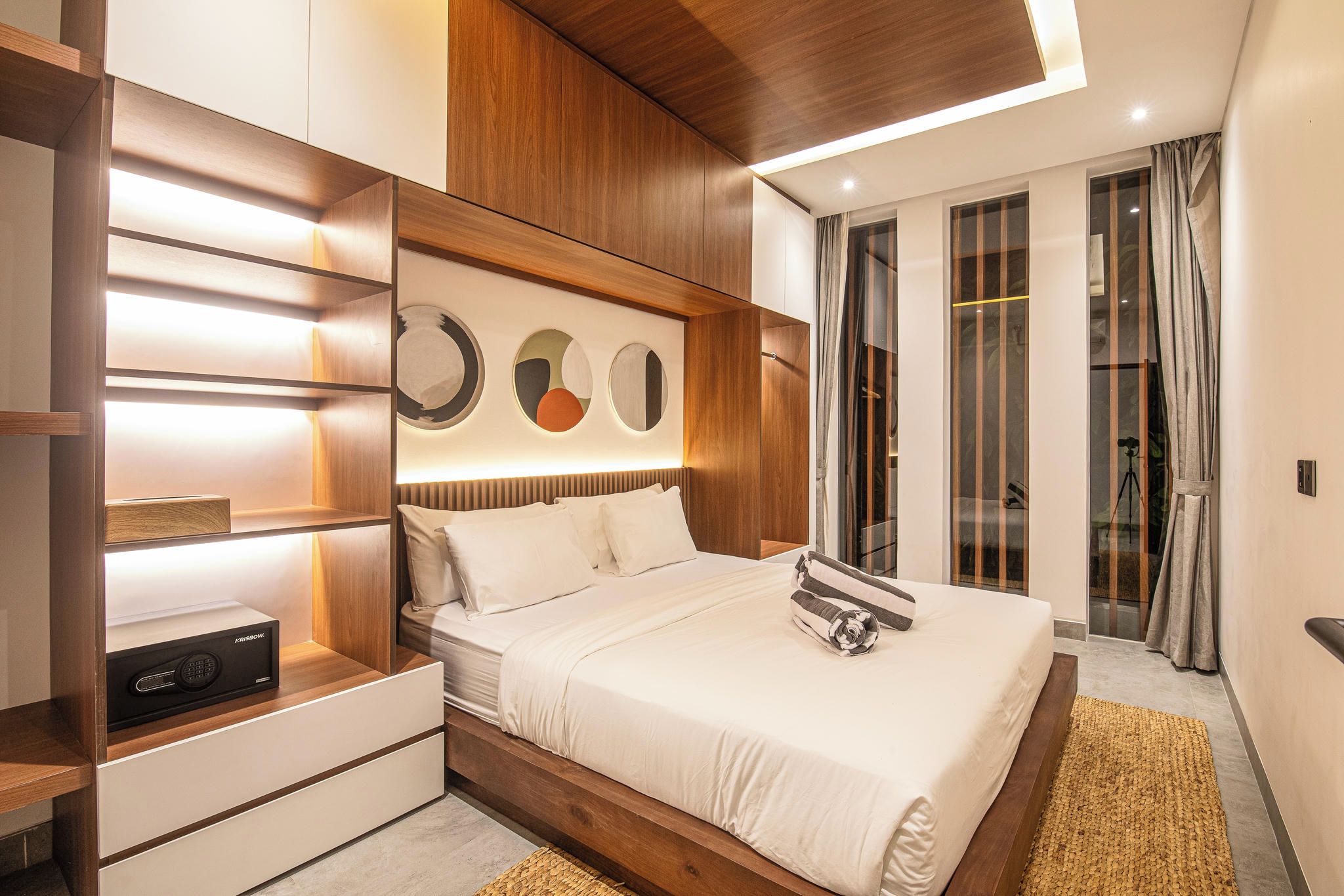 A modern bedroom with a wooden headboard, a bedside table with a radio, a large bed with a white comforter, and a view of the outdoors through a glass door.