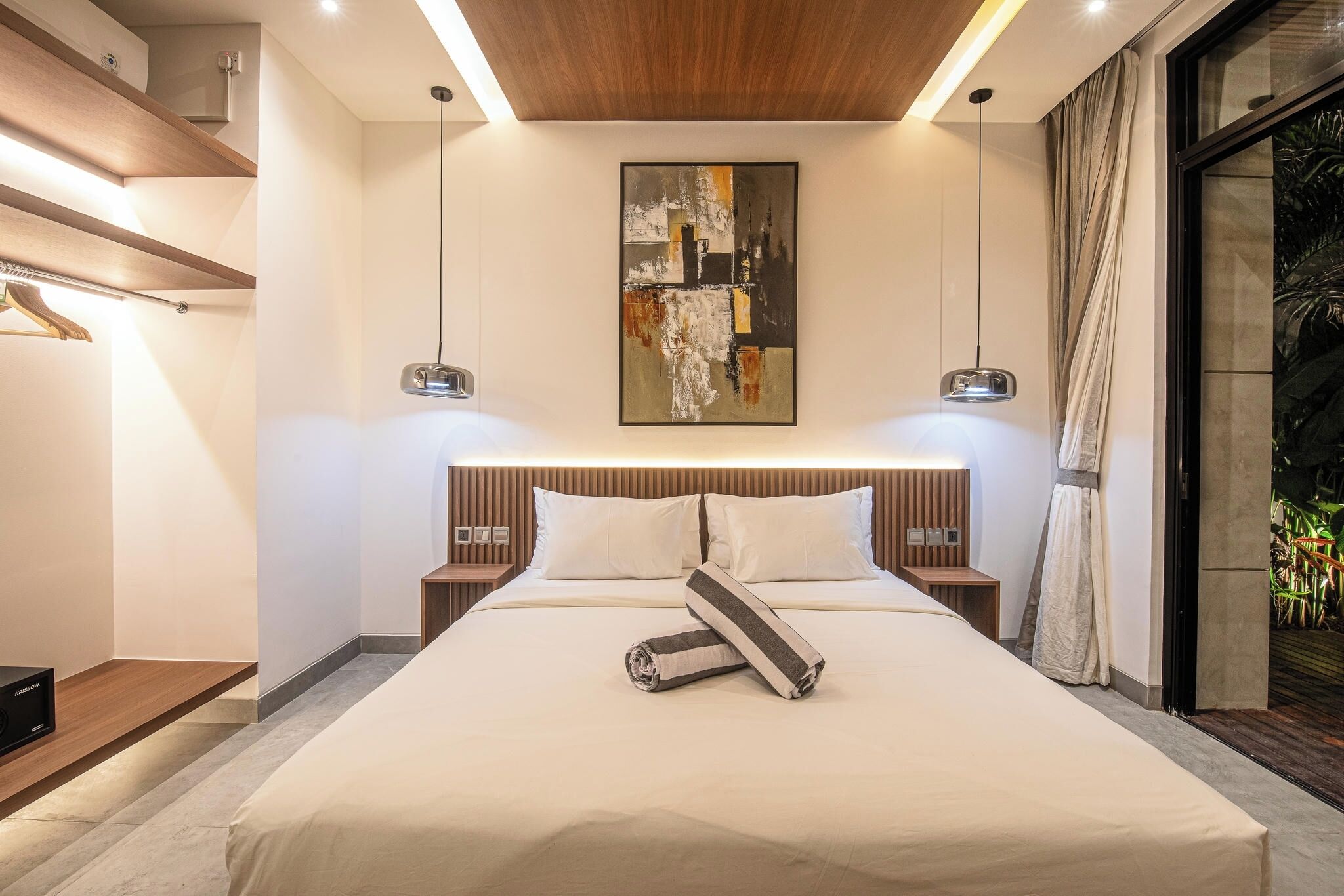 Un hotel moderno con una cama grande, cabecera de madera, mesitas de noche, un cuadro abstracto en la pared y vistas a zonas verdes a través de una ventana.