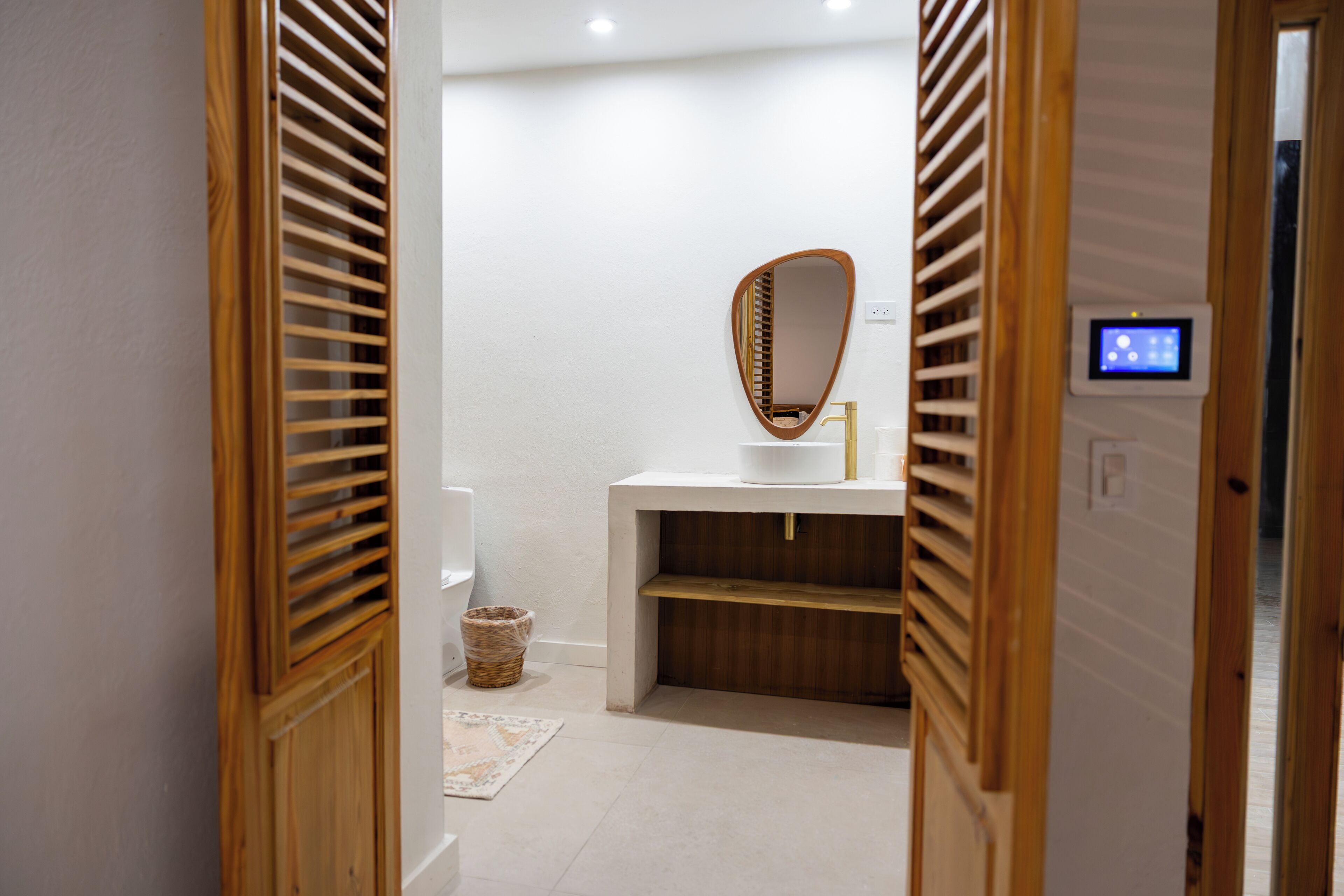A hallway leading to a bathroom with a wooden door, a white sink, a mirror, and a control panel on the wall.