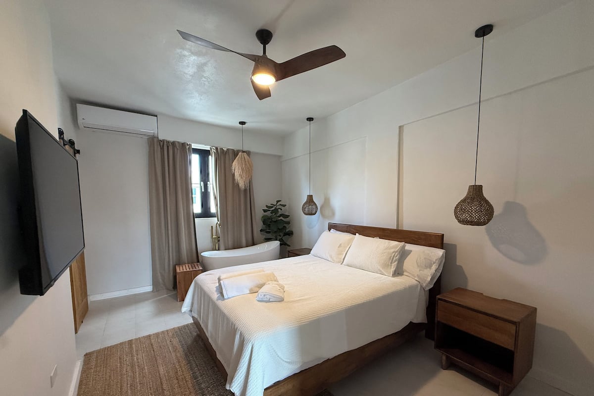 Un dormitorio con cama, mesita de noche, televisor, ventilador de techo y ventana con cortinas.