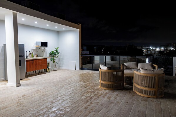 Eine Dachterrasse mit Küchenbereich, Sitzgelegenheiten und Blick auf die beleuchtete Stadt.