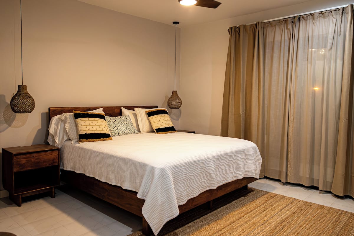 Una cama bien tendida con una colcha blanca y almohadones estampados, un cabecero de madera y dos lámparas colgantes.