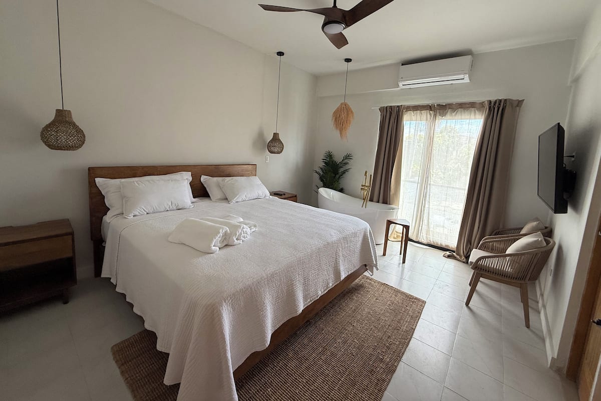 Un dormitorio con una cama grande, un ventilador de techo, un televisor y una ventana con cortinas.