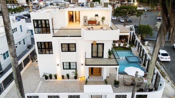 Un edificio moderno de varios pisos con piscina en la azotea, un área de estar al aire libre techada y una cocina visible.