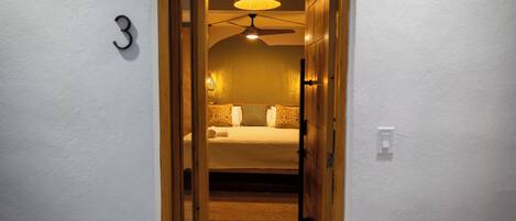 A narrow hallway leading to a bedroom with a bed, a ceiling fan, and a lamp.