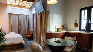 Una habitación de hotel moderna con cama, una mesa de comedor de vidrio, sofá, televisor y zona de cocina.