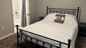 Ein Schlafzimmer mit Bett, Nachttisch, geschlossener Tür und einem durch eine offene Tür einsehbaren Badezimmer.