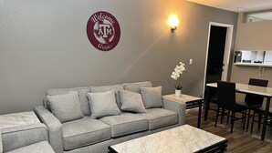 Ein modernes Wohnzimmer mit einer grauen Eckcouch, einem Marmortisch und einem Essbereich mit Tisch und Stühlen. Eine wandmontierte Leuchte und ein Willkommensschild sind sichtbar.