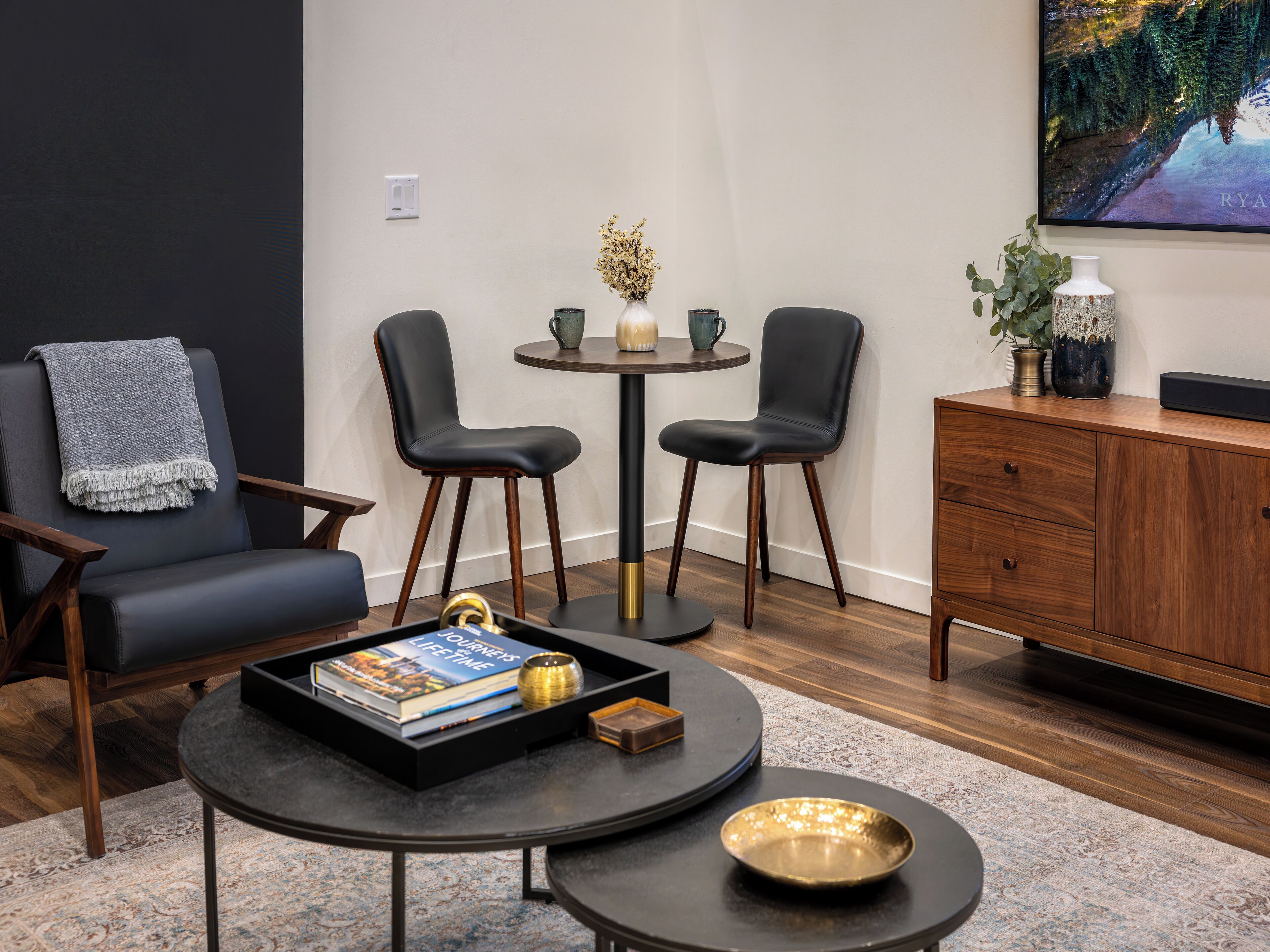 A modern living room with a dark leather sofa, two black chairs, a small round table with a vase and a book, and a wooden cabinet.
