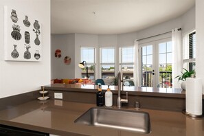 Eine moderne Küche mit einer Edelstahl-Spüle, einer Arbeitsplatte mit einer Handseifenflasche und einem Fenster mit Blick auf einen Balkon.
