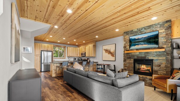 Een moderne woonkamer met een grijze modulaire bank, houten plafond, stenen open haard en een keuken op de achtergrond.