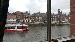 Une rivière avec un bateau rouge et blanc, des bâtiments le long de la rive et le clocher d’une église en arrière-plan.