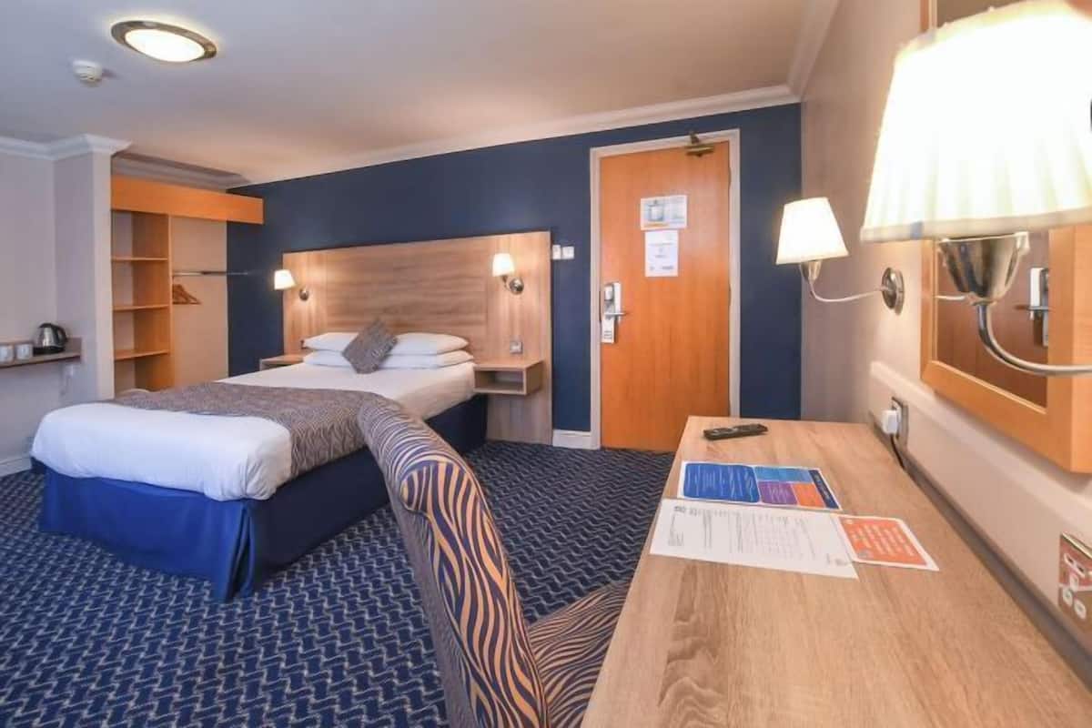 Une chambre d’hôtel avec un lit, une table de chevet, une lampe et un bureau avec des papiers.