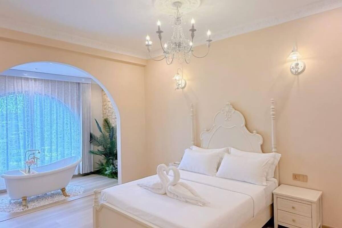 A bedroom with a white bed, a chandelier, a white nightstand, and a bathtub.