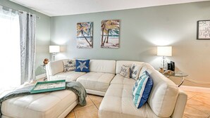 A living room with a beige sofa, two wall-mounted paintings, a glass coffee table, a lamp, and a side table with a jar. - Steps to Sand: Condo w/ Pool Access in Cocoa Beach (Cocoa Beach)