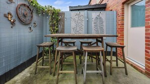 En terrasse med et træbord og fire barstole, et dekorativt ur samt et hegn i metal med bladmotiver.