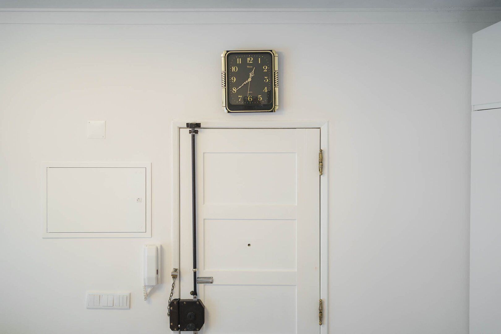 Una puerta blanca con un reloj negro encima, un interruptor de luz y un termostato en la pared.