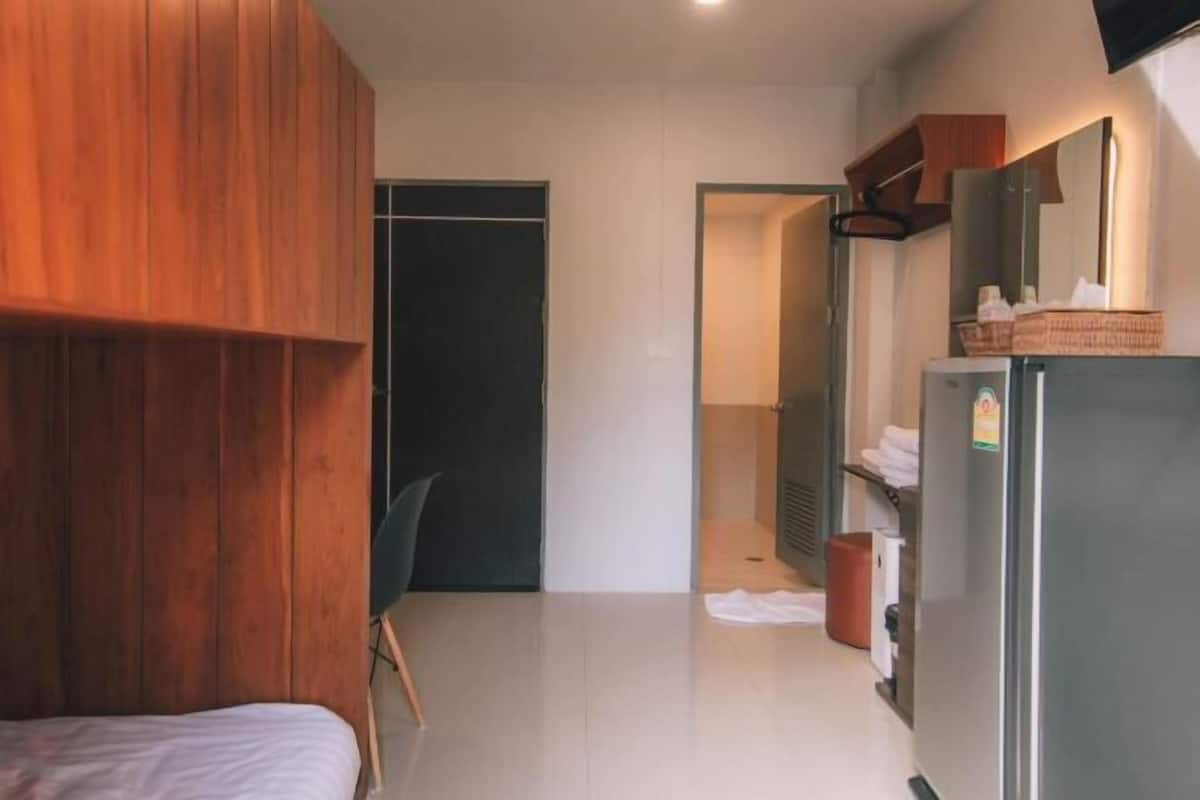 A modern hotel room with a wooden wardrobe, a bed, a small desk, a refrigerator, and a bathroom door.