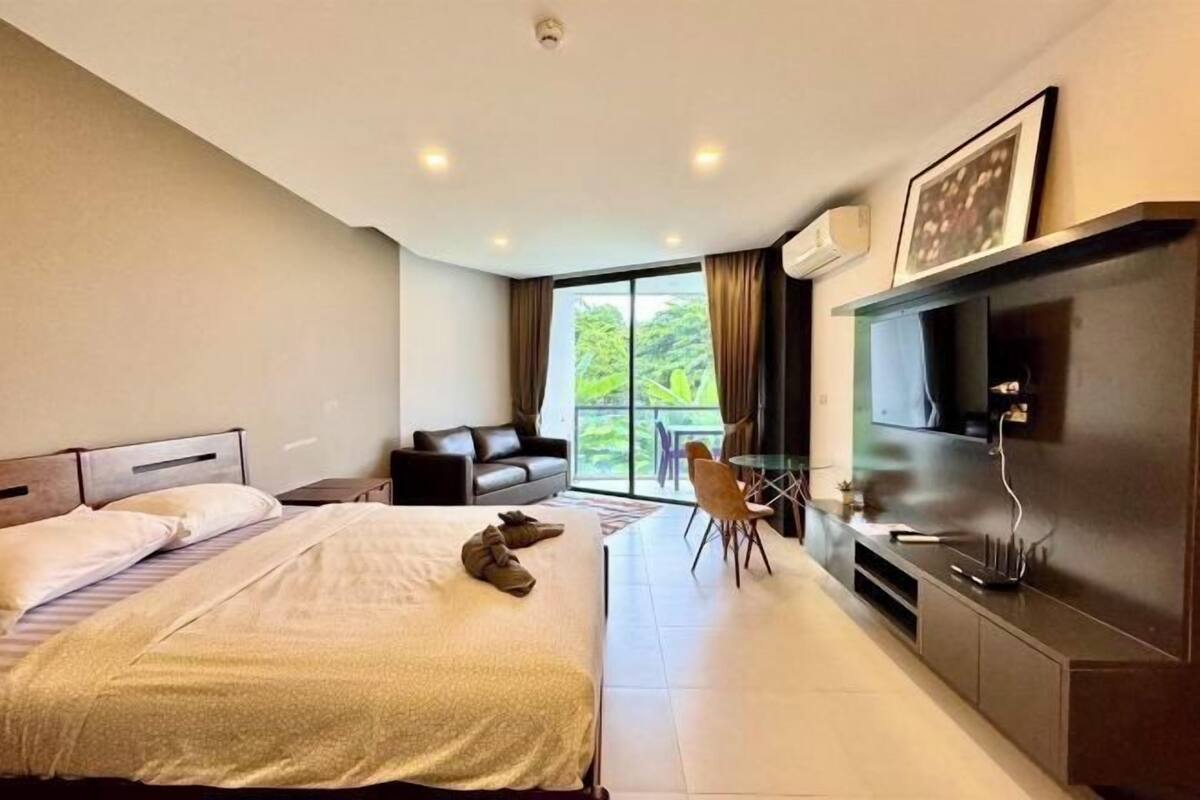 A hotel room with a bed, a sofa, a TV, and a balcony with a view of greenery.
