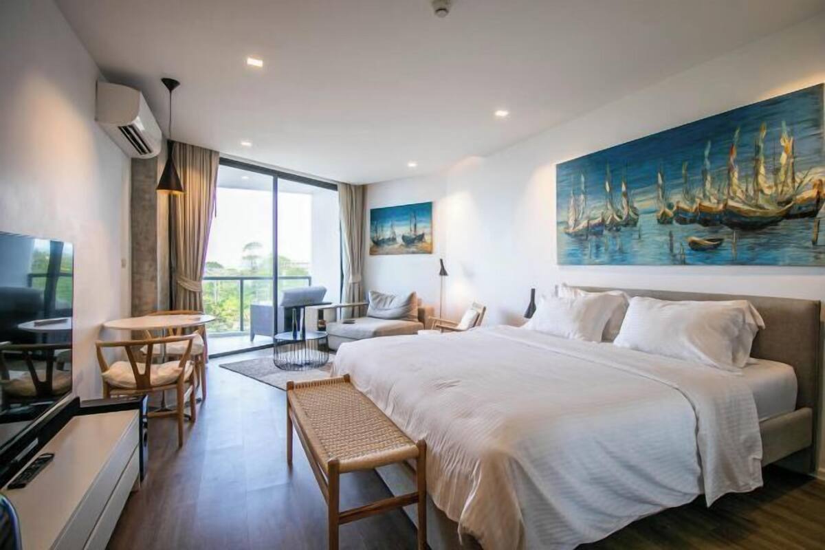 A modern hotel room with a large bed, a dining area, and a painting of boats on the wall.