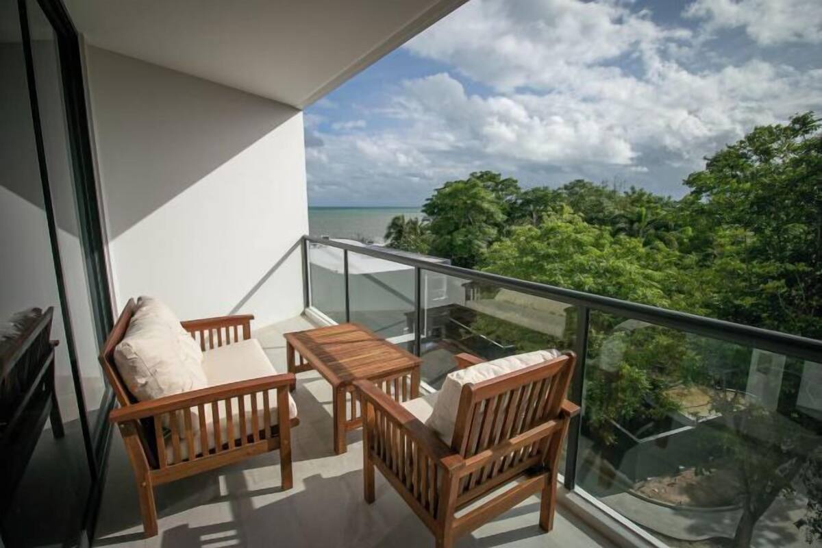 A balcony with wooden furniture and a glass railing overlooking a lush green landscape and a body of water.