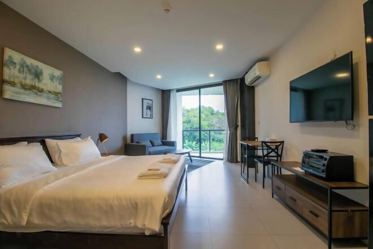 A hotel room with a bed, a desk, a chair, a TV, and a window with a view of greenery.