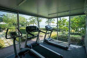 A gym with treadmills and an elliptical machine, large windows with a view of palm trees and a body of water. - Escape Condominium Beachfront Suites - Mae Phim (Klaeng)