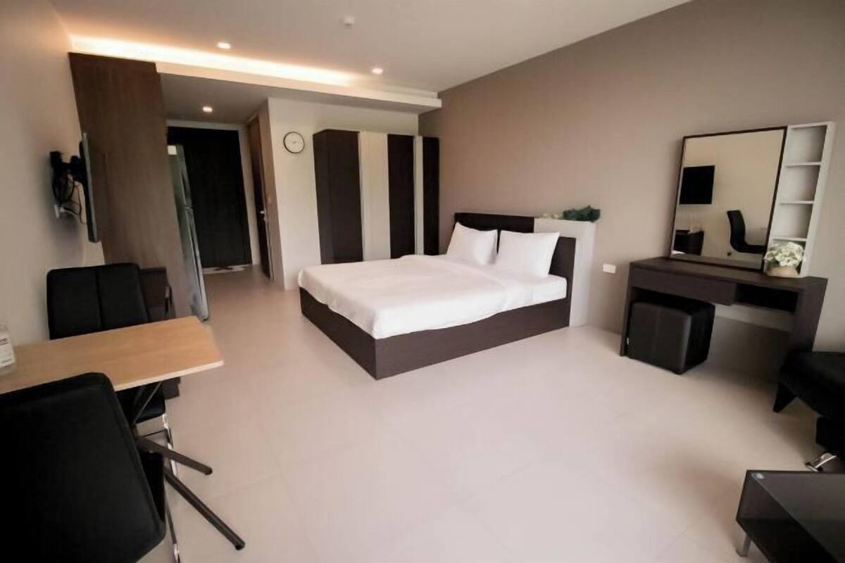 A modern hotel room with a bed, a desk, a chair, a mirror, and a small cabinet.