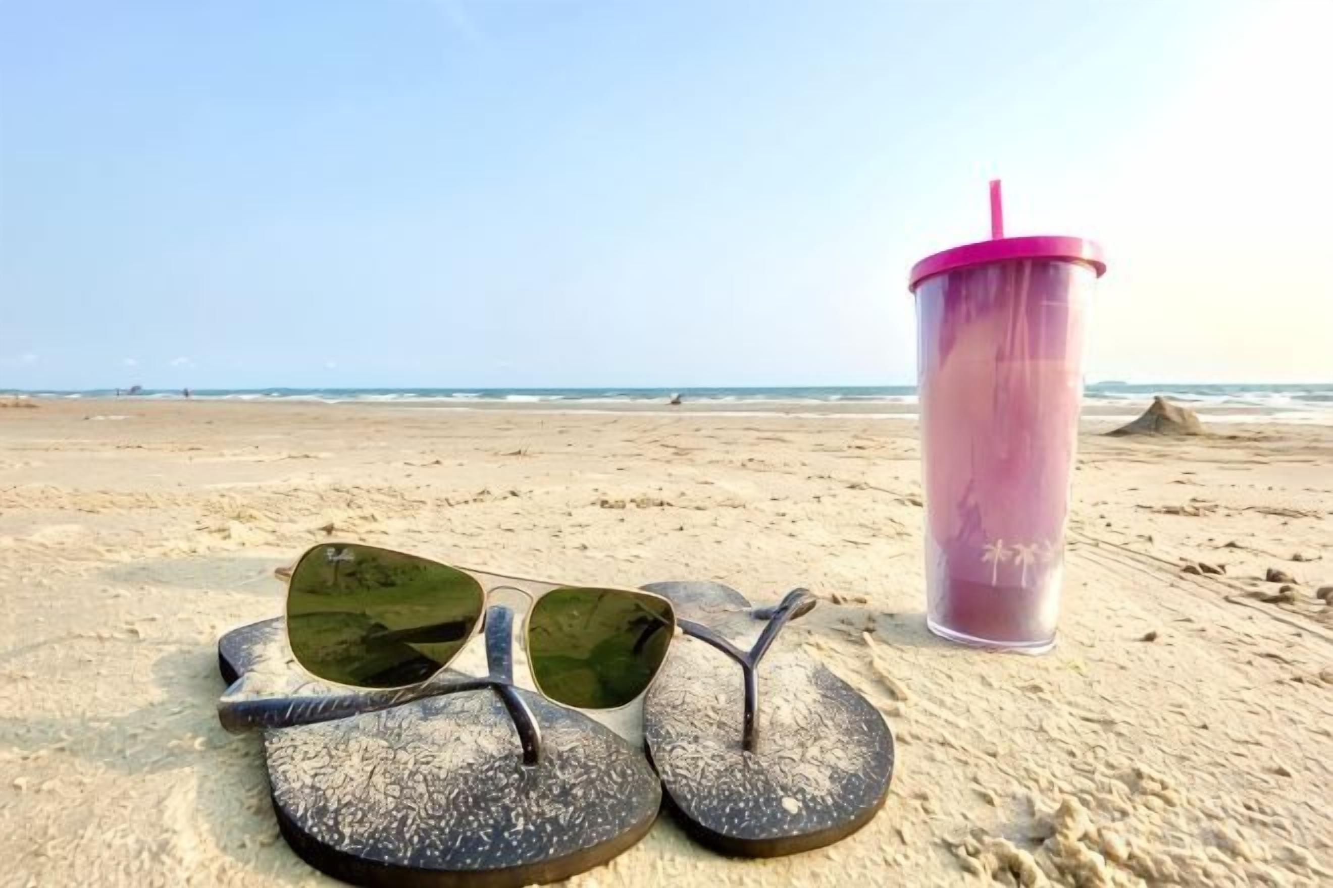 Chanclas y un vaso en una playa arenosa.