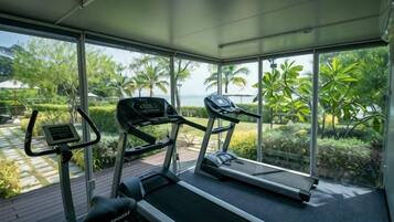 Un gimnasio con cintas de correr y una bicicleta estática, amplias ventanas con vistas a palmeras y una masa de agua.