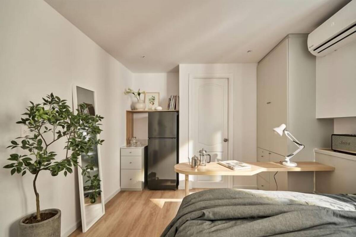 Un dormitorio pequeño con cama, escritorio, silla, planta en maceta, espejo, armario y una kitchenette compacta.