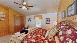Quarto com paredes de madeira, cama com roupa de cama floral, ventoinha no teto e janela com persianas.