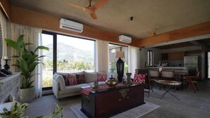 Ein großzügiger Wohnbereich mit einer Couch, einem Couchtisch und einem Essbereich mit Stühlen. Große Fenster bieten einen Blick ins Freie.