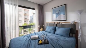 Une chambre d’hôtel avec un lit, une table de chevet, une lampe, une fenêtre avec des rideaux, un tableau encadré au mur et une vue sur les bâtiments à l’extérieur.