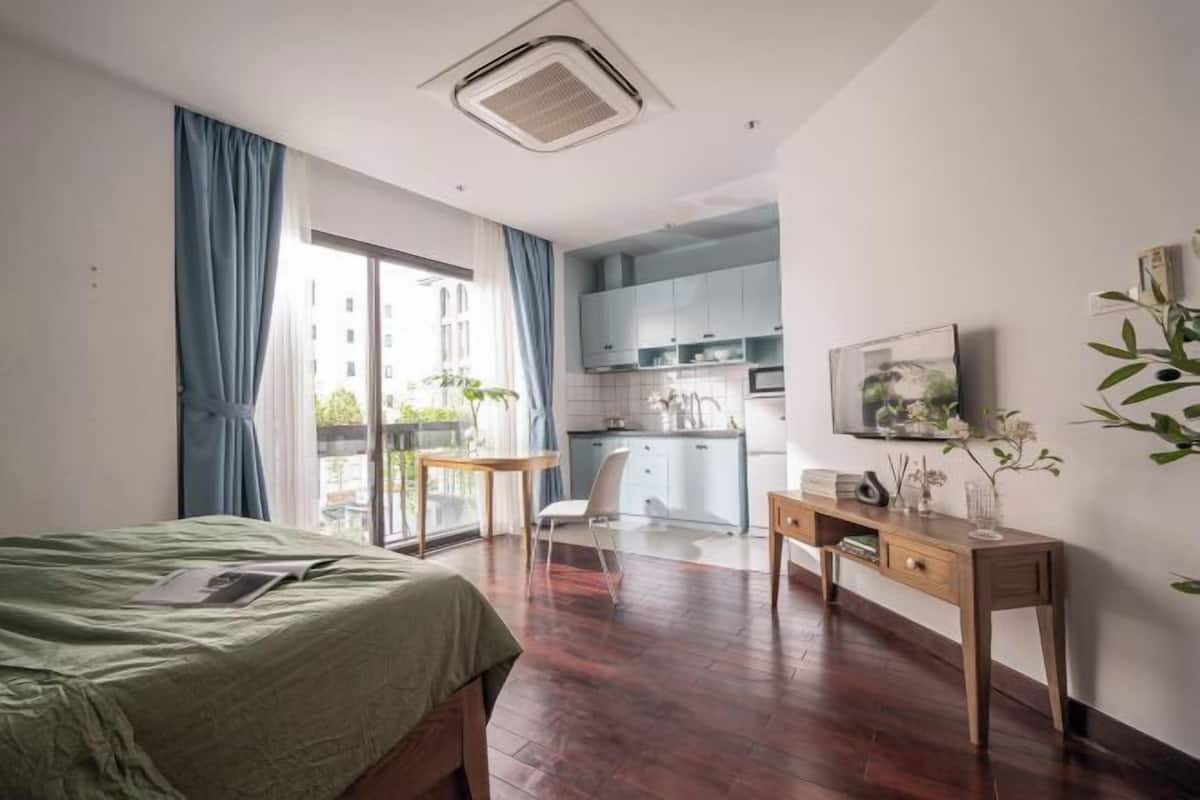 A bedroom with a bed, a desk, a chair, a window with curtains, a kitchen area, and a mounted picture.