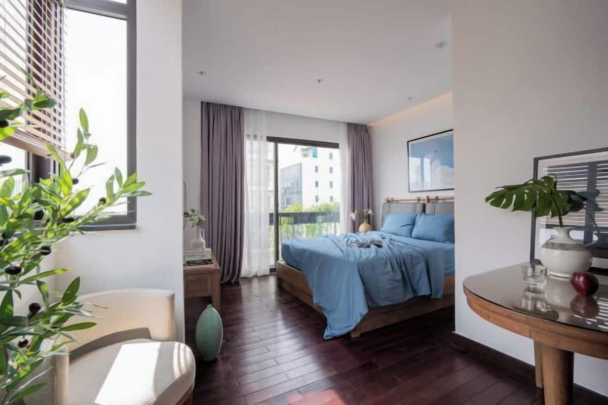 A modern hotel room with a bed, a desk, a chair, and a balcony with a view of buildings.