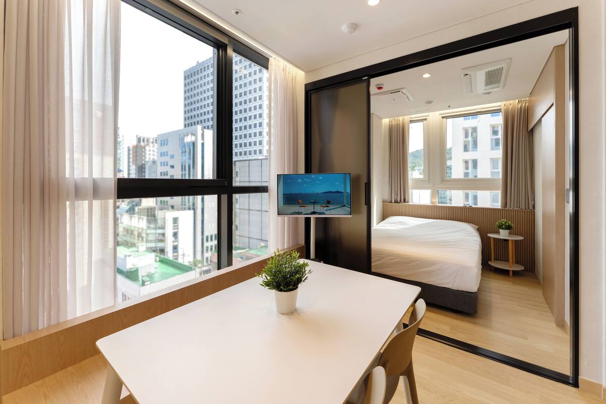 Habitación de hotel moderna con un ventanal grande, una cama, una mesita con una planta y un televisor en la pared.