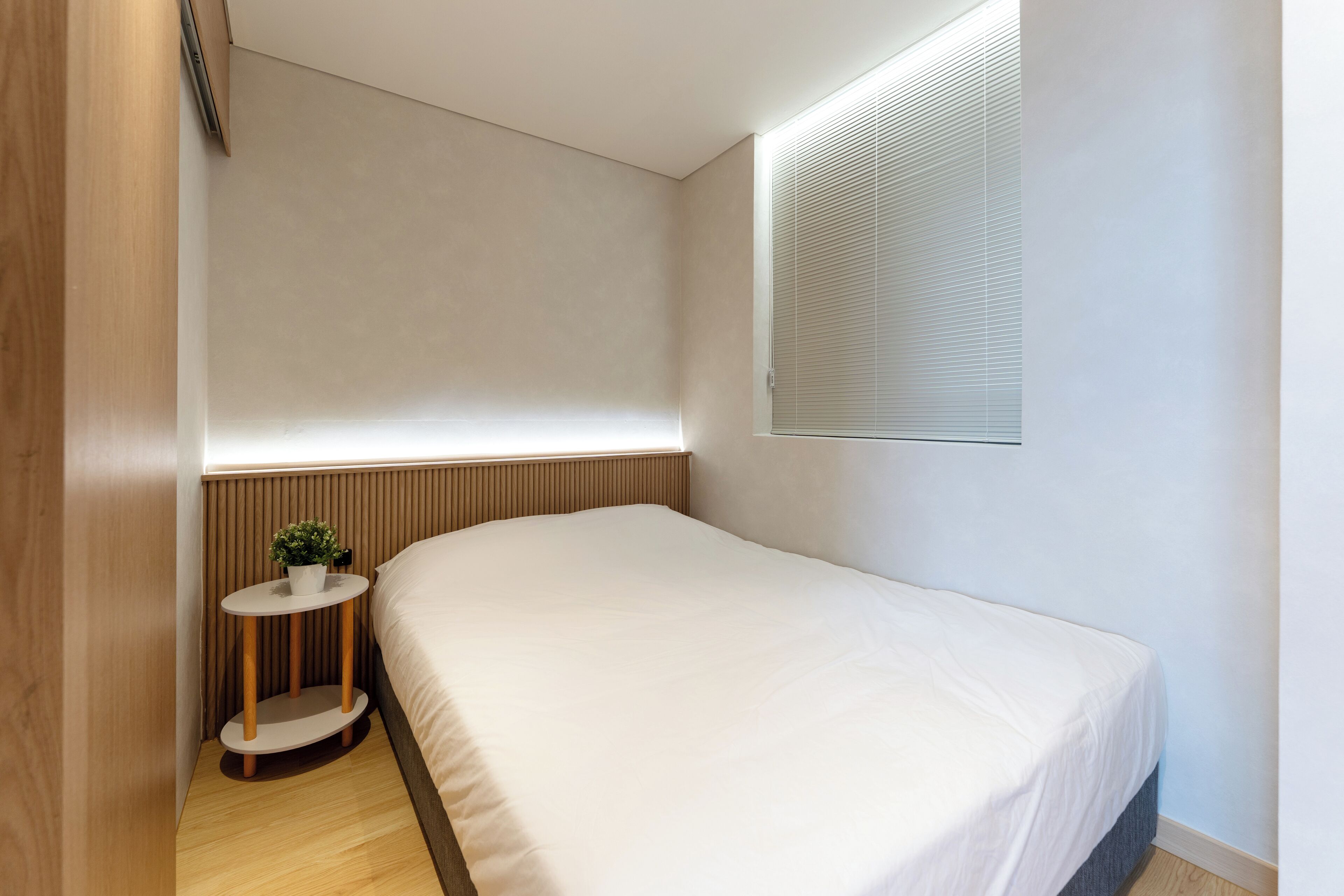 Een moderne slaapkamer met een groot bed, een klein nachtkastje met een plant en een raam met zonwering.