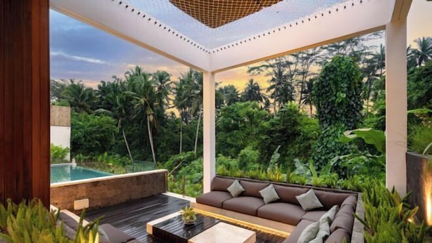 Una zona relax esterna moderna con divano e tavolino, circondata da vegetazione rigogliosa e vista su una piscina.