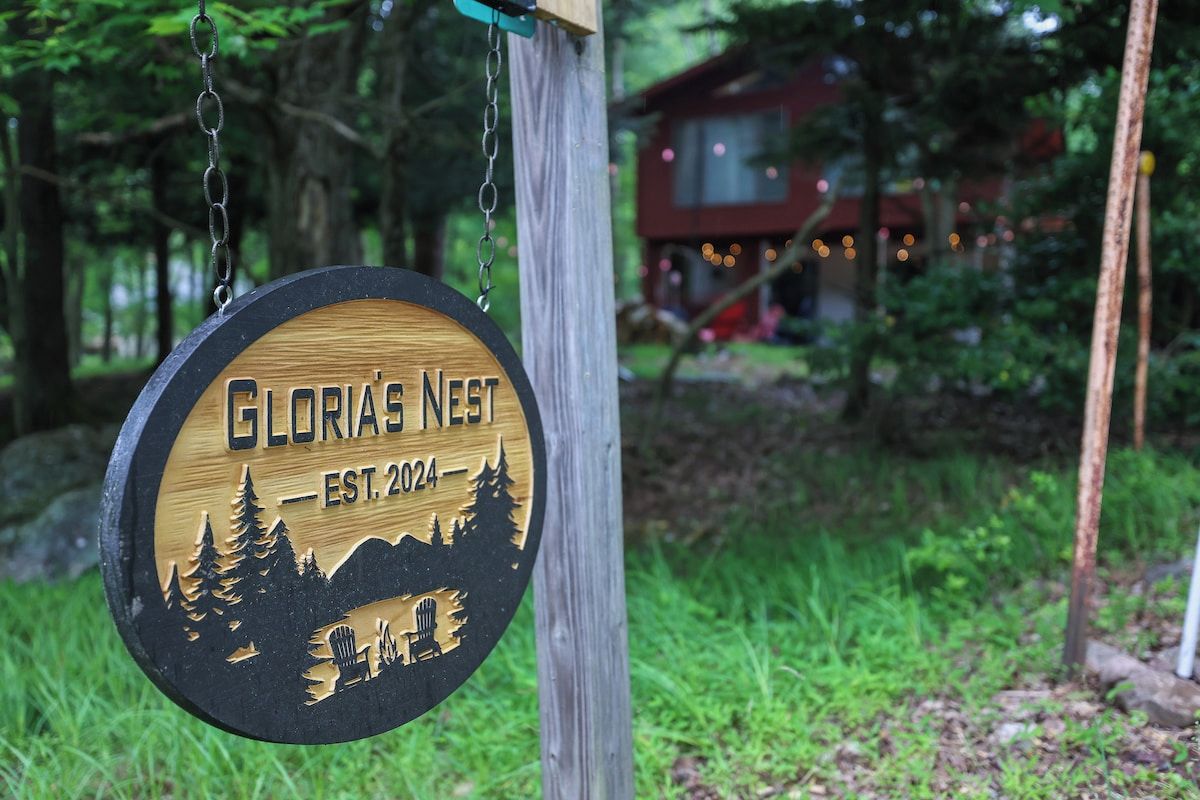 A wooden sign with the text 'Gloria's Nest EST. 2024' and a silhouette of a cabin surrounded by trees.