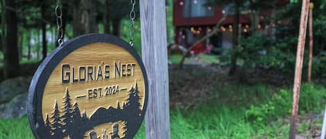 A wooden sign with the text 'Gloria's Nest EST. 2024' and a silhouette of a cabin surrounded by trees.