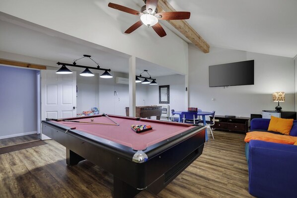 Une table de billard, un ventilateur de plafond, un téléviseur à écran plat et un canapé bleu se trouvent dans une pièce dont le sol est recouvert de parquet.