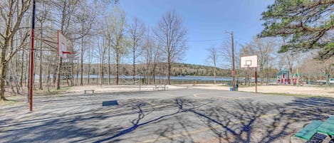 Un terrain de basket en plein air avec un banc, des arbres et une aire de jeux en arrière-plan.