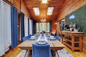 Une salle à manger en bois avec une longue table dressée pour six personnes, bordée de rideaux bleus et adossée à un mur vert.