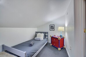 Un lit simple avec un matelas gris et un oreiller blanc, un meuble de rangement rouge sur roulettes, une lampe fixée au mur et un tableau encadré accroché au mur.