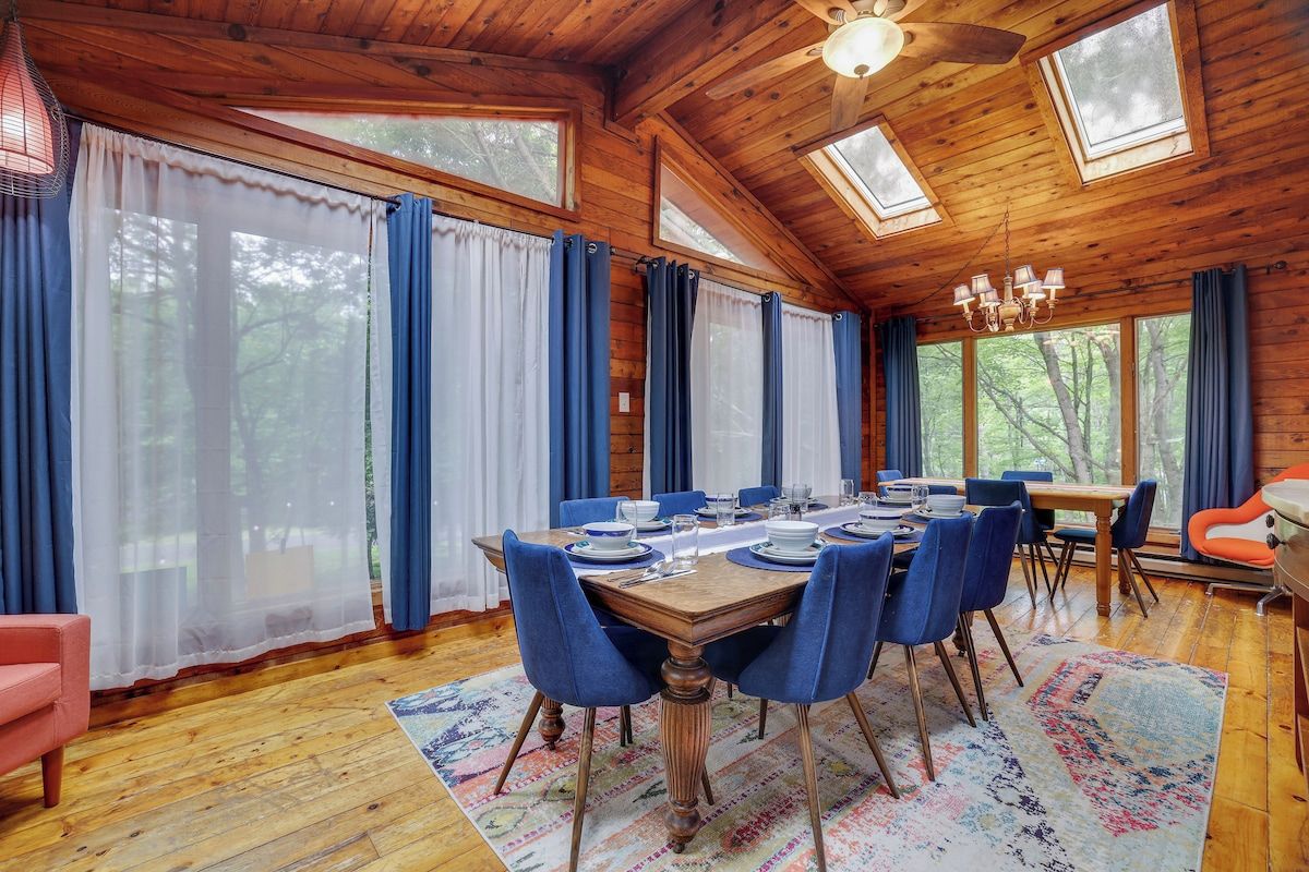 Uma sala de jantar com uma mesa de madeira posta para seis pessoas, rodeada por cadeiras azuis, em uma cabana com paredes e teto inclinados de madeira.
