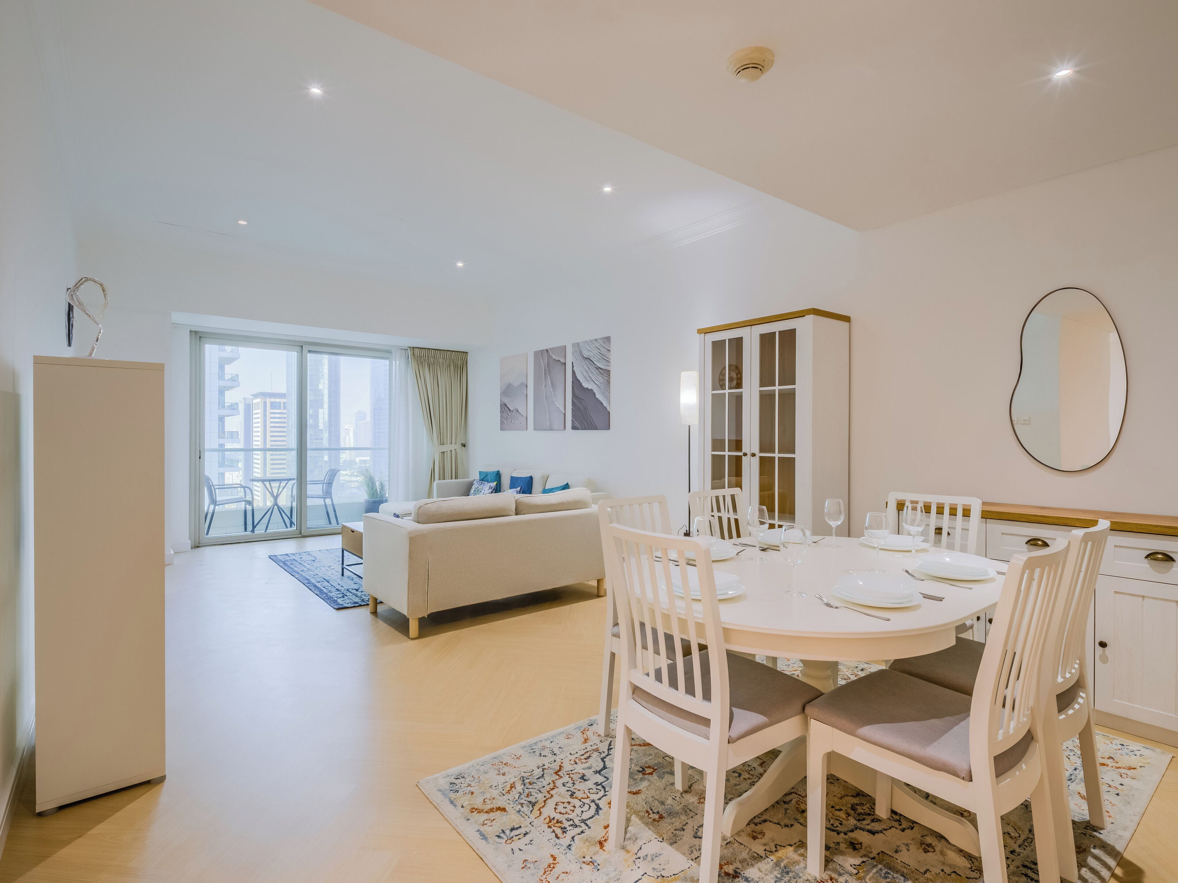 A modern living room with a sofa, dining table, and chairs. There is a sliding glass door leading to a balcony with outdoor furniture.