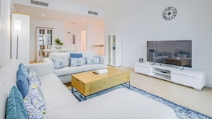 Uma sala de estar moderna com um sofá branco, uma mesa de centro e uma TV sobre um suporte branco. Ao fundo, há uma área de jantar com uma mesa e cadeiras.